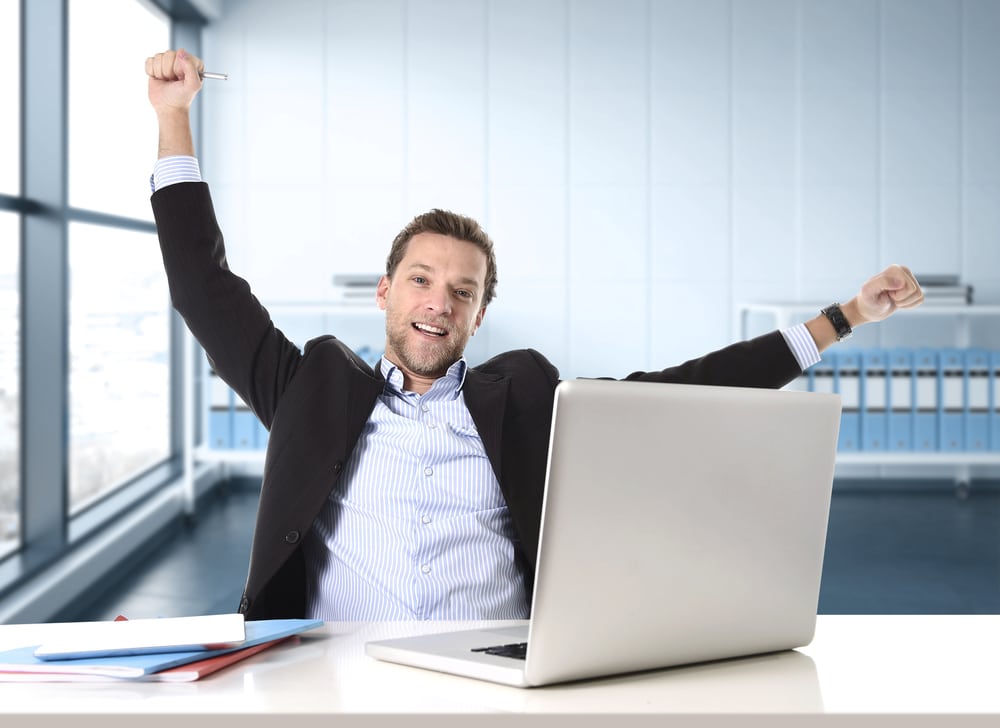 Young, enthusiastic businessman celebrating a career milestone at his computer desk, smiling in a modern office with a window.